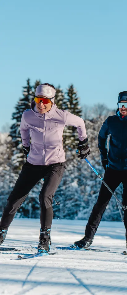 Two people cross-country skiing on a snowy trail with alpine mountains in the background