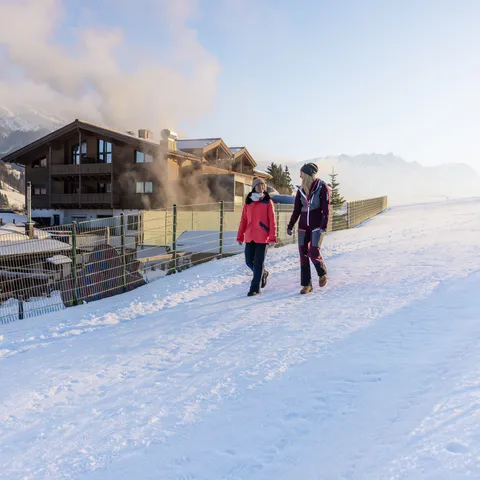 Zwei Frauen spazieren auf einem verschneiten Weg neben Alpenchalets mit Bergblick