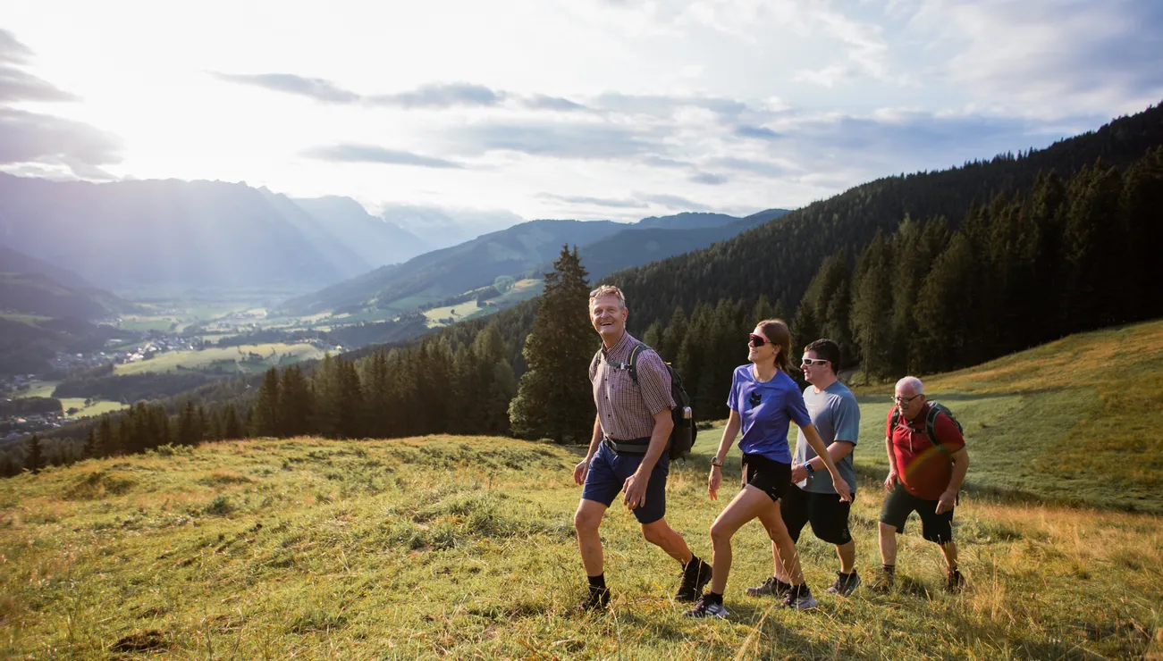 Gruppe wandert bergauf durch Almwiese mit Bergtal und Wald im Hintergrund