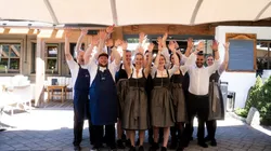Smiling restaurant staff in traditional Bavarian attire raising hands outside a rustic venue