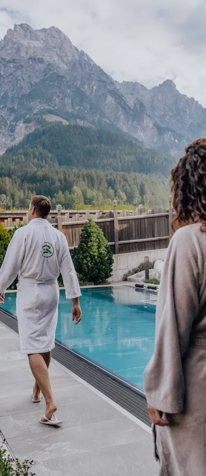 Paar im Bademantel geht am Außenpool eines Wellnesshotels in den Alpen entlang