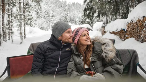 Happy couple sitting in a horse sleigh surrounded by snowy forest and stacked firewood