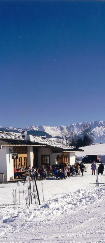 Skihütte mit Menschen vor der Tür an verschneitem Hang in den Alpen bei blauem Himmel