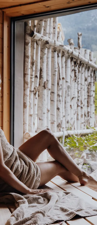 Frau entspannt in einer Holzsauna mit Blick auf Berge und Garten