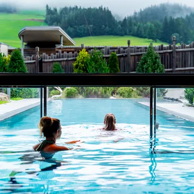 Zwei Frauen schwimmen in einem Infinity-Pool mit Blick auf grüne Hügellandschaft
