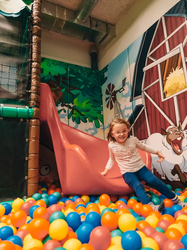 Smiling girl sliding into indoor ball pit with colorful farm-themed wall art