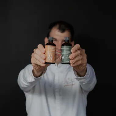 Mann in weißem Hemd hält zwei Flaschen fermentierte Garum-Saucen vor dunklem Hintergrund