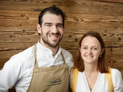 Two smiling people in traditional attire standing in front of wooden panel wall