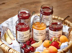 Verschiedene Marmeladengläser mit Obst in einem geflochtenen Korb auf Holztisch
