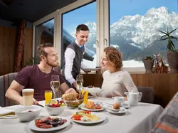 Paar genießt Hotel-Frühstück mit Bergblick, während Kellner Essen serviert